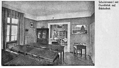 Schulzimmer I mit Schulbänken, kleinem Lehrerpult unter einer Abbildung des Völkerschlachtdenkmals sowie Durchgang zur Schulbibliothek