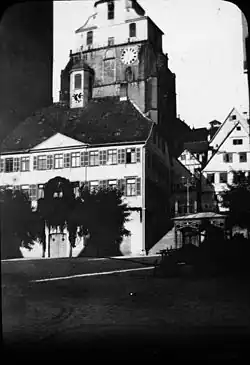An einem Marktplatz mit Kopfsteinpflaster stehen einige große Leiterwagen, ein Rathaus mit kleinem Uhren- und Glockenturm, rechts davon ein über eine Treppe erreichbarer höher gelegener Platz mit mehreren Fachwerkhäusern. Dahinter etwas höher an einem Berg eine große Kirche mit massivem Kirchturm, die Spitze ist wegen des Bildrandes nicht zu sehen.