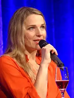 Tanja Wedhorn mit langen Haaren spricht in ein schnurloses Stabmikrofon, welches sie mit der rechten Hand hält. Sie trägt ein orange-rotes Oberteil und eine Kette mit einem Anhänger. Sie sitzt vor einem blauen Vorhang und vor ihr seht ein halb geleertes Glas mit Rotwein.