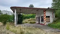 Eingewachsene und beschmierte Tankstelle. In den Fensterlöchern befinden sich Bretter. Der weiße Putz platzt ab.