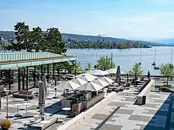 Man sieht von oben auf die Terrasse mit Sonnenschirmen, links das Restaurant, hinten im Bild den Zürichsee sowie die Berge.