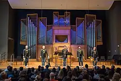Fünf männliche Personen mittleren und ähnlichen Alters mit unterschiedlichen Musikinstrumenten in der Hand auf einer Bühne vor dem Publikum stehend von vorne in gleicher Kleidung - blaue Jeans, schwarzes T-Shirt mit gleichem Symbol in gelb, dunklem Jacket und weißen Schuhen in Farbe.