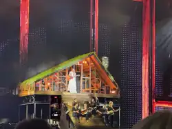 Taylor Swift steht in einem weißen Kleid an einem Standmikrofon. Auf der Treppe vor ihr sitzt ihre Band.