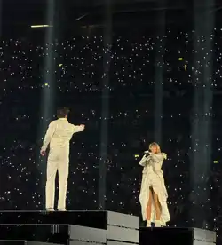 Taylor Swift und Jan Ravnik stehen in weißen Klamotten auf Hydraulik-Plattformen auf einer Bühne. Beide zeigen aufeinander. Swift steht der Kamera zugewandt und singt zeitgleich in ein Mikrofon, das sie in der anderen Hand hält.