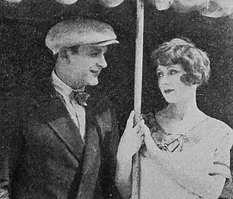 Standbild aus dem amerikanischen Liebesdrama „The Prince of Pep“ (1925) mit Richard Talmadge und Nola Luxford, auf Seite 67 des Exhibitors Herald vom 12. Dezember 1925. Das Bild zeigt zwei Personen, die sich unter einem Baldachin oder Sonnenschirm befinden. Beide stehen nebeneinander. Links befindet sich Richard Talmadge. Er trägt einen klassischen Anzug, ergänzt durch eine Krawatte und eine Schirmmütze. Rechts steht Nola Luxford und trägt ein elegantes Kleid mit einem dekorativen Kragen. Beide Hände halten den Stab des Baldachins oder Sonnenschirms, der über ihnen gespannt ist.
