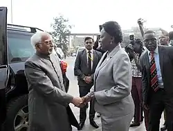 Ein älterer Mann und eine Frau schütteln sich die Hand. Im Hintergrund sind Fotografen und Sicherheitsleute.