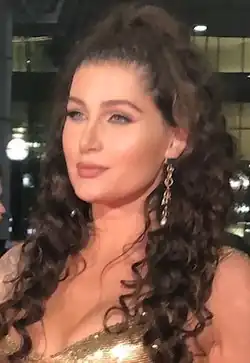 Porträtfoto von Trace Lysette. Sie trägt ein goldfarbenes Kleid, am linken Ohr eine goldene Kette mit einem silbernen, steinförmigen Ohrring und ihre braunen, schulterlangen Haare als nach unten stehende Locken. Im Hintergrund sind das Gesicht einer anderen Person, runde Straßenlampen und ein Gebäude mit mehreren Fenstern zu sehen.