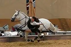 Eine Frau scheint lächelnd im Spagat neben einem Pferd in vollem Galopp zu schweben. Bei genauem Hinsehen sieht man, dass sie sich mit einem Arm am Sattel festhält und die ausgestreckten Beine (eines nach hinten, eines nach vorne) in speziellen Steigbügeln stecken.