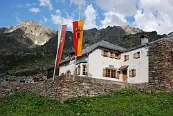 Schrägaufnahme auf eine Alpenhütte; im Hauptgebäude verputzt, sonst roher Stein; vor der Hütte zwei Masten mit Fahnen; im Hintergrund ein aufsteigendes Bergmassiv aus rohen Fels