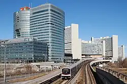 Zug der Type V auf der U1 vor der Skyline der Donau City; im Hintergrund die Station Kaisermühlen – Vienna International Centre