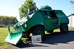 Grün lackierter, mit Räumschild, Blaulicht und Schriftzug POLIZEI versehener gepanzerter Wagen von demselben Typ wie auf dem Bild weiter oben, aufgenommen von links vorne. Die in der Fahrzeugmitte befindliche seitliche Schwenktür ist offen. Das Fahrzeug ist ein Ausstellungsstück; vor ihm ist einer Tafel mit Erläuterungen montiert. Zu erkennen sind zwei Überschriften: „Thyssen Rheinstahl UR 416 Geschützter Polizei-Sonderwagen“ und „Thyssen Rheinstahl UR 416 im weltweiten Einsatz“.