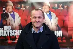 Maximilian Brückner steht vor einem Filmplakat bei der Österreichpremiere von dem Film Und Äktschn! im Gartenbaukino in Wien. Auf dem Plakat ist ein Mann abgebildet, der eine Kamera hält. Er trägt eine beige Weste über einem blauen Hemd. Der Hintergrund des Plakats zeigt rote Theatersitze, die eine cineastische Atmosphäre schaffen. Der Titel des Films lautet in großen weißen Buchstaben: „...UND ÄKTSCHN!“. Unter dem Titel sind mehrere Namen in kleinerer Schrift aufgelistet, darunter „KARL MARKOVICS“, „MICHAEL OSTROWSKI“ und „DIRK STERMANN“.