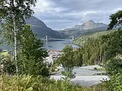Im Vordergrund ein Dorf im Grünen, im Hintergrund eine den Fjord überspannende Hängebrücke.
