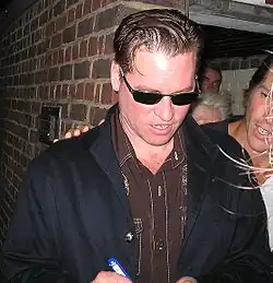 Val Kilmer (2005) steht vor einer Ziegelwand. Er trägt einen dunklen Mantel über einem braunen Hemd, das sichtbare Nähte und Knöpfe aufweist. In der rechten Hand hält er einen blauen Stift und scheint etwas zu unterschreiben. Am rechten Rand des Bildes ist teilweise eine andere Person erkennbar, die einen schwarzen Mantel über einem weißen Hemd trägt. Der Ort wirkt wie ein Außengelände, möglicherweise in der Nähe eines Gebäudeeingangs.