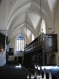 Frontale Farbfotografie aus dem Innenraum einer Kirche mit dunklen Bänken und dunkler Empore an der rechten Seite. Links hängen an den Wänden und Pfeilern Gemälde. An der hinteren Wand ist ein farbiges Kirchenfenster und von dem weißen Gewölbe hängen Kronleuchter.