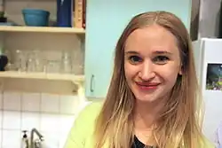 Vera Vitali Junge Frau mit langen, blonden Haaren und lächelnden Gesicht, die in einer modernen Küche steh lächelndem Gesicht, trägt ein helles Oberteil, steht in einer Küche. Die Haare fallen locker über die Schultern, und sie trägt ein hellgrünes Oberteil über einem schwarzen Shirt.Die Küche ist mit hellen Schränken ausgestattet, die glatte Fronten haben. Eine weiße, geflieste Rückwand sorgt für eine saubere und helle Atmosphäre. Auf den Regalen stehen verschiedene Gegenstände wie Gläser und Schüsseln.Links unten im Bild ist ein Waschbecken mit einer Armatur sichtbar. Im Hintergrund sieht man einen Kühlschrank, an dessen Tür ein kleines Bild oder Magnet angebracht ist.Die gesamte Szene vermittelt den Eindruck eines gemütlichen und funktionalen Küchenbereichs in einer privaten Wohnung.