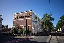 Ein Bild des Verwaltungsgebäudes der Privatbrauerei Herrenhausen. Dunkelbraune Backsteinfassade mit gelben Schriftzug "Brauerei Herrenhausen" in Frakturschrift und dem Logo der Brauerei, einem steigenden weißen Pferd auf rotem Grund.