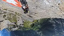Kletterer am Leukerbad-Klettersteig auf Höhe der ikonischen Metalltafel mit der Schweizer Flagge, aus etwa 2320 m Blick herab auf das darunter gelegene Leukerbad