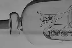 Schwarz-Weiß Bild des oberen Drittels einer liegenden Flasche, in der sich ein gläsernes Schiff befindet. Vom Schiff ist der als Drachenkopf geformte Bug zu sehen.