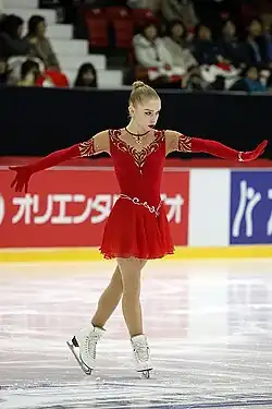 Viveca Lindfors steht in der Pose einer Balletttänzerin in der Mitte der Eisfläche. Sie trägt ein rotes Kleid und rote, über die Ellenbogen reichende Handschuhe, ihre blonden Haare sind zu einem Dutt gebunden. Im Hintergrund japanische Bandenwerbung und ein Ausschnitt der Publikumstribüne.