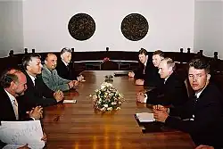 Mehrere Männer sitzen sich an einer hölzernen Tafel gegenüber. Auf der Tafel steht ein Blumenstrauß.