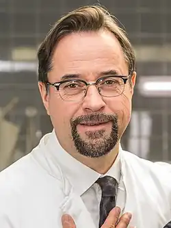 Jan Josef Liefers lächelt freundlich in die Kamera und trägt einen weißen Laborkittel, und eine gemusterte Krawatte. Er hat hellbraunes Haar und hält mit einer Hand den Laborkittel fest. Im Hintergrund ist eine Laborumgebung zu erkennen, mit gefliesten Wänden und einigen Geräten, die schemenhaft sichtbar sind. Das Foto enstandbei den WDR-Dreharbeiten zu Tatort Münster „Lakritz“ in der Rolle des Prof. Dr. Dr. Karl-Friedrich Boerne 2019.