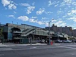 Hochbahnhof in dunkel-grünlichem Erscheinungsbild mit kleinteiligen Rasterflächen aus leicht spiegelnden Fenstern sowie organisch anmutenden netzförmigen Überdachungen von Zugangstreppenhäusern. Unten Kreuzung von breiten Hauptstraßen mit Personen und Autos an Verkehrsampel, oben blauer Himmel mit Wölkchen, im Hintergrund Großwohnhäuser mit Backsteinverblendung.