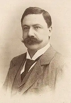 Wolfram Zingerle mit langem, dunklem Schnurrbart, schwarzer Krawatte und Anzug