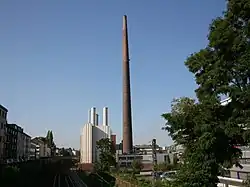 Blick über eine Eisenbahnstrecke auf einen teilweise metallverkleideten Gebäudekomplex in städtischer Umgebung. Das Foto wird in der Mitte von dem riesigen Backsteinschornstein dominiert. Links daneben ein deutlich niedrigerer, kastenförmiger, fensterloser Baublock mit heller Verkleidung und mehreren niedrigen, metallenen Schornsteinen über dem Dach.