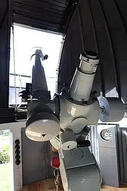 Ein großes Linsenteleskop ragt zur Dachluke hinaus. Im Innern ist es dunkel, Tageslicht dringt durch die Dachluke.