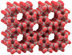 Eine Computergrafik aus grauen und roten Kugeln. Die Kugeln sind teilweise ineinander geschoben und bauen so eine räumliche Struktur auf. Graue Kugeln sind paarweise durch jeweils eine rote Kugel verbunden. In der Struktur sind fünf zylindrische Hohlräume, um die herum sich das Netzwerk der Kugeln legt. Die Hohlräume sind untereinander symmetrisch angeordnet und ihr Durchmesser ist in etwa genau so breit wie der Abstand zwischen zwei Hohlräumen.