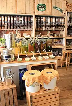 Der Verkaufsbereich in einen niederländischen Unverpacktladen. 6 Glasbehälter gefüllt mit verschiedenen Lebensmittelölen sind in der Mitte aufgereiht, darunter Olivenöl, Semsamöl und Balsamico. Dahinter befinden sich, in einem hölzernen Wandregal befestigt, hohe transparente Glaszylinder mit trockenen Lebensmitteln wie Getreideflocken und mit Schokolade überzogenen Leckereien.