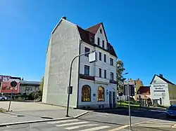 einzelnes Haus einer ehemaligen Häuserzeile, einspurig mit Zebrastreifen, im Hintergrund Bushaltestelle und Reklame