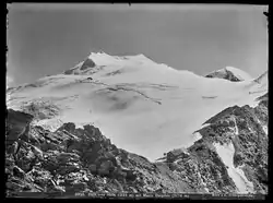 Schwarz-Weiß Fotografie eines stark vergletscherten Berggipfels. Am unteren Bildrand zieht sich ein Felsband durch das Bild, etwas rechts von der Mitte sieht man eine Berghütte.