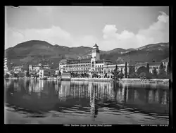 Schwarz-Weiß Fotografie, auf der man eine Häuserzeile sieht, die sich im Wasser eines Sees spiegelt. Im Hintergrund bewaldete Hügel.