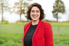 Person mit kurzen braunen Haaren und runder Brille, lächelnd in einem roten Blazer über schwarzer Bluse. Im Hintergrund eine grüne Wiese mit gelben Blumen und Bäumen. Aufnahme im Freien bei Tageslicht.